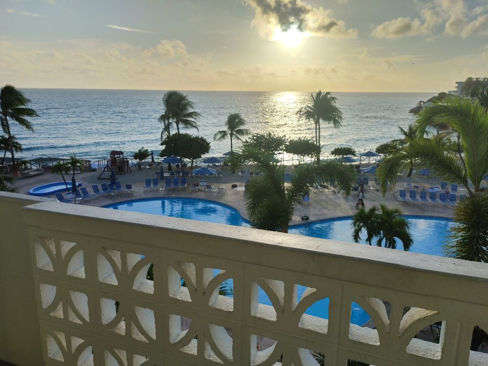 The sun is setting over a view from a balcony on St Maarten, showing a lovely pool below, just before sand and palm trees of the beach beyond. The sun reflecting on the water makes it a little difficult to tell, but the ocean is that famous clear blue of the Caribbean.