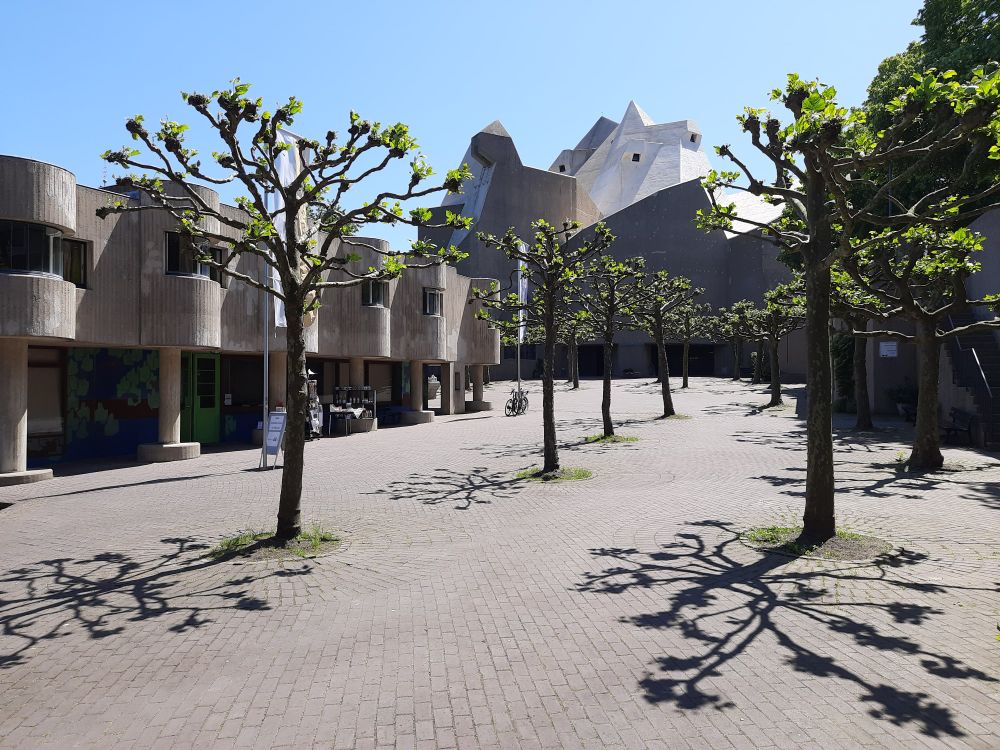 Im Vordergrund Kopfweiden, die auf einen gepflasterten Platz Schatten werfen. Im Hintergrund eine moderne Kirche.