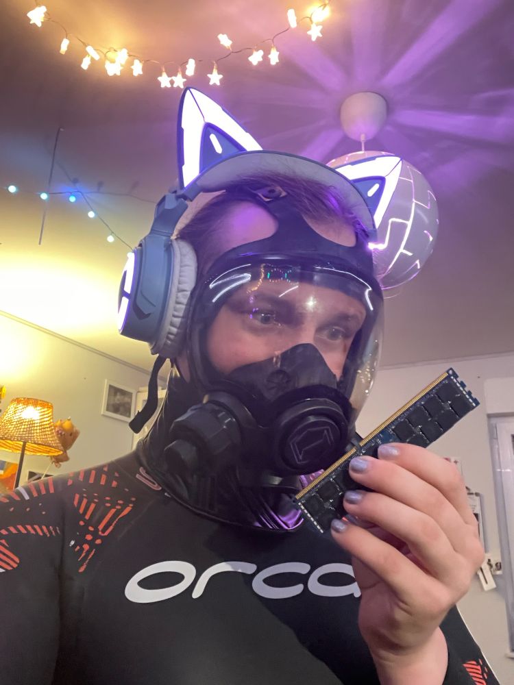 Hexdrone 4703 in wetsuit, gasmask and cat ear headset looking at a RAM Stick its holding