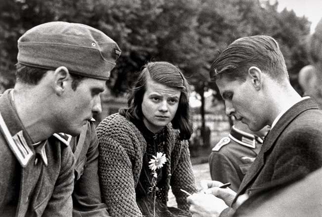 1942 or 1943 black and white photo of three young Germans sitting near trees in what appears to be a park:  a young man in a military uniform, a dark-haired young woman in civilian clothes, and a young man in civilian clothes. They are Hans Scholl, his sister Sophie Scholl, and their friend Christoph Probst, members of the White Rose Society who protested against the Nazi government. The Nazis executed them by guillotine on 22 February 1943.