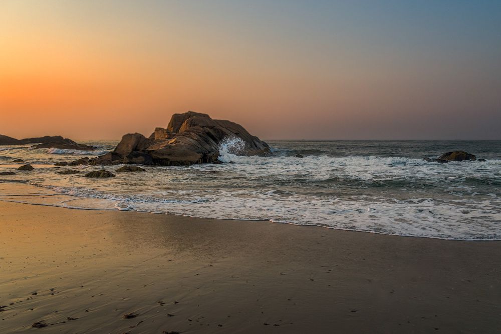 Beach at sunrise. Sun is lightly hitting a large rock on the left. The sky is orange and blue. Small waves. 