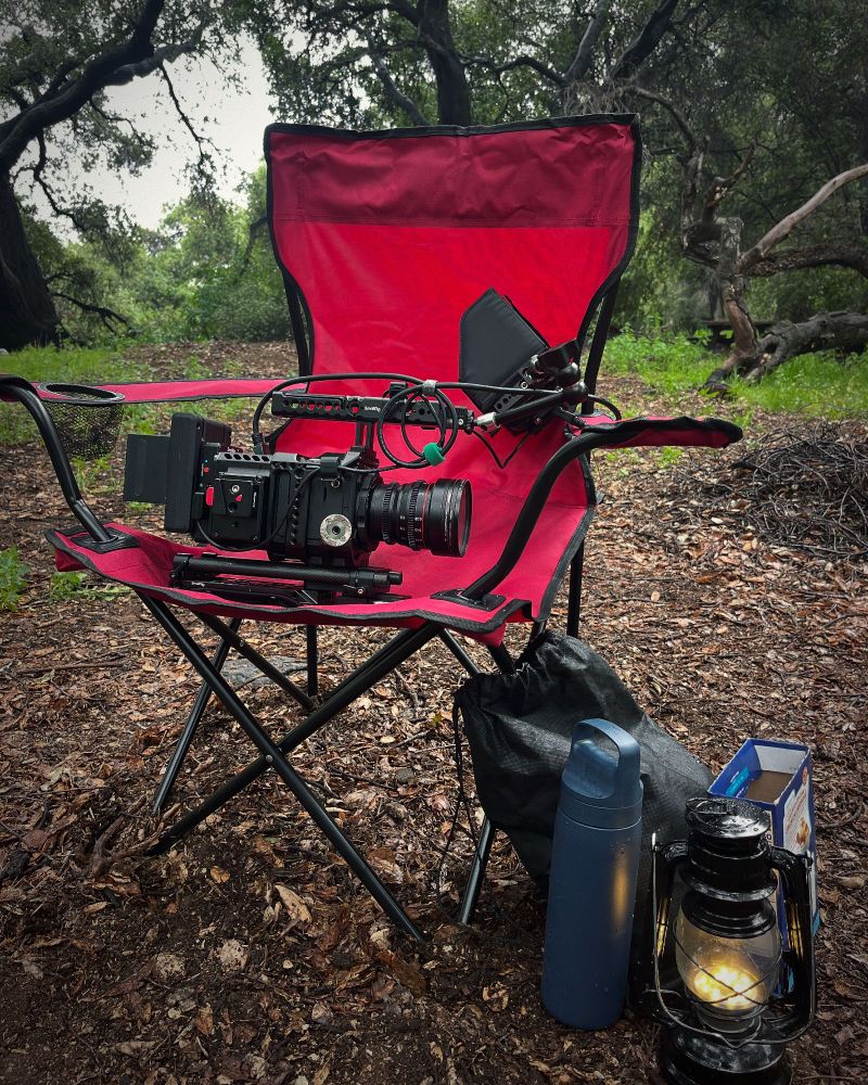 A red, foldable camping chair in the middle of the woods. A high-tech camera is resting on the chair's seat. Next to the chair on the dirt and leaf-covered ground is a lantern, a black satchel, and a thermos. 