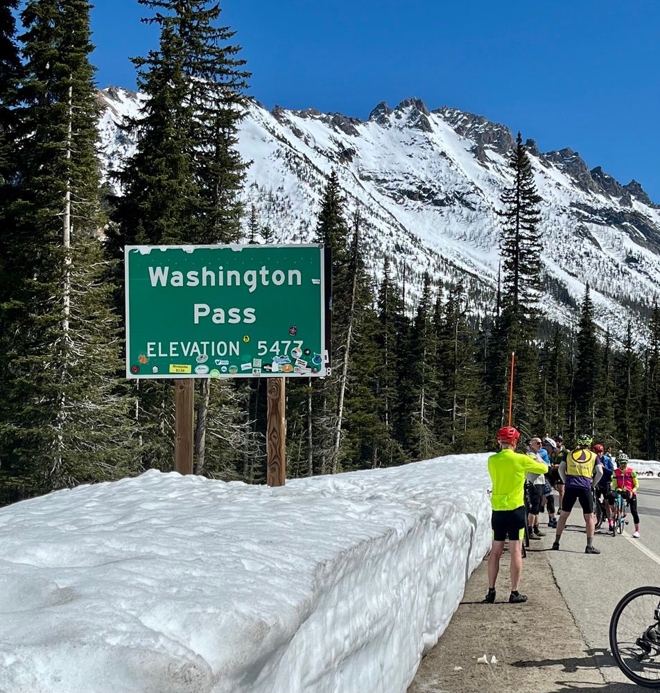 Green road sign reads “Washington Pass; Elevation 5473.” In the background are evergreen trees and snowy mountains. A bank of snow covers the side of the roadway. A group of cyclists stand talking and taking photos next to the snow. 