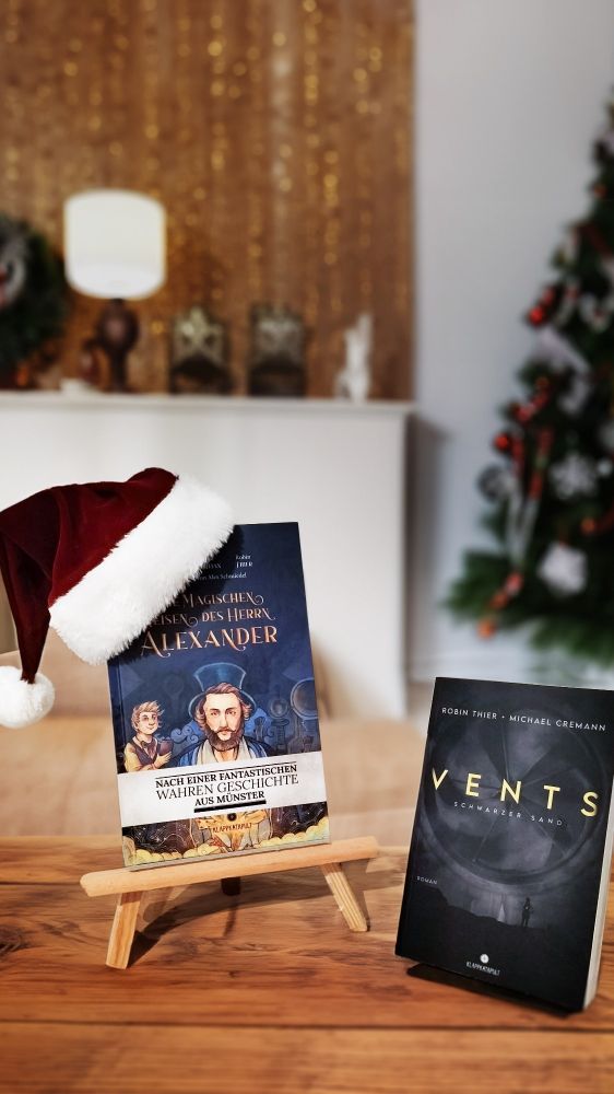 Ob weihnachtlichem Ambiente die Bücher "die magischen Reisen des Herrn Alexander" und "Vents". Das linke Buch trägt eine Weihnachtsmütze.