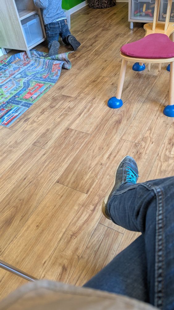 Auf dem Bild sieht man einen Innenraum, der wie ein Kinderzimmer oder Spielbereich wirkt.

Links kniet ein kleines Kind auf dem Boden und schaut oder greift in ein offenes Regal. Vor ihm liegt ein bunter Spielteppich mit Straßen- und Stadtmotiven, der etwas verrutscht ist.

Der Boden besteht aus warm wirkendem Holz oder Laminat mit sichtbarer Maserung. Rechts steht ein kleiner Kinderstuhl mit blauen runden Gummikappen an den Stuhlbeinen und einem roten Sitzpolster.

Im Vordergrund sieht man einen Teil einer sitzenden Person – ein Bein in dunkler Jeans und ein abgenutzter Sneaker mit türkisen Schnürsenkeln.

Der Raum wirkt ordentlich, kindgerecht und einladend.