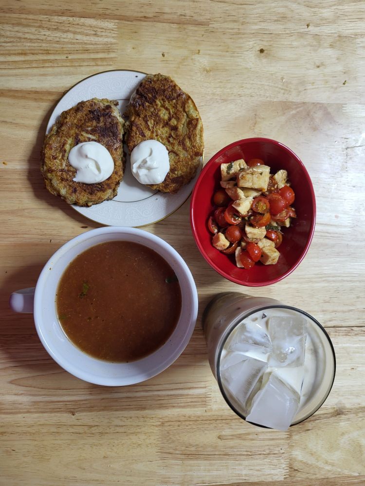Photo of a lunch spread. Two well browned fritters on a small china plate, each with a dollop of sour cream, sit next to a small red bowl of halved cherry tomatoes and cubed fresh mozzarella, darkened with balsamic vinegar. They are accompanied by a ceramic mug bowl full of a dark red brown broth, all with a tall glass of ice swimming in dairy. 
