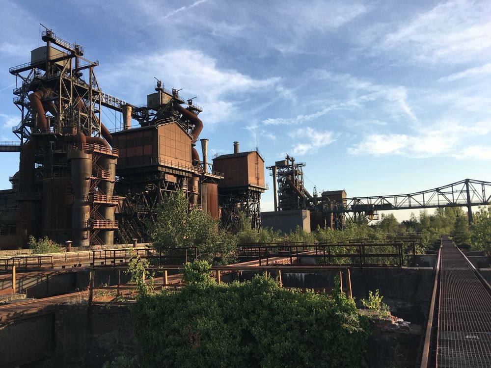 Image shows a walkway across the former storage pits, now filled with greenery, with sunlit blast furnaces behind