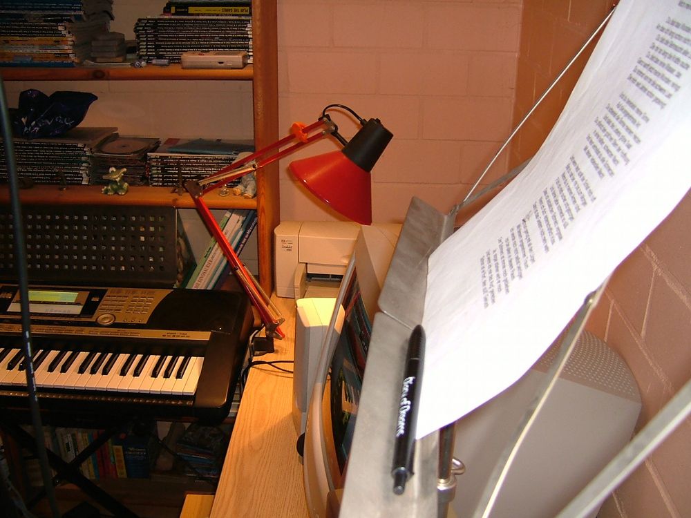 Side view of Dominik Sonders' first improvised home studio, simple desk with printer and old-school computer screen, a note stand with lyrics, a music keyboard, in the background a shelf with stuffed animals, video game magazines, a Gameboy etc.
