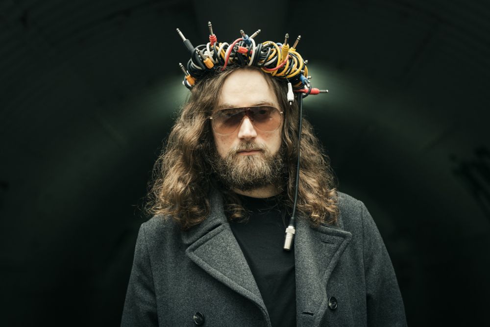 A photo of Dominik Sonders alias Sidetrack Walker: A long-haired, bearded man wearing a crown of audio cables, standing in a pedestrian tunnel.