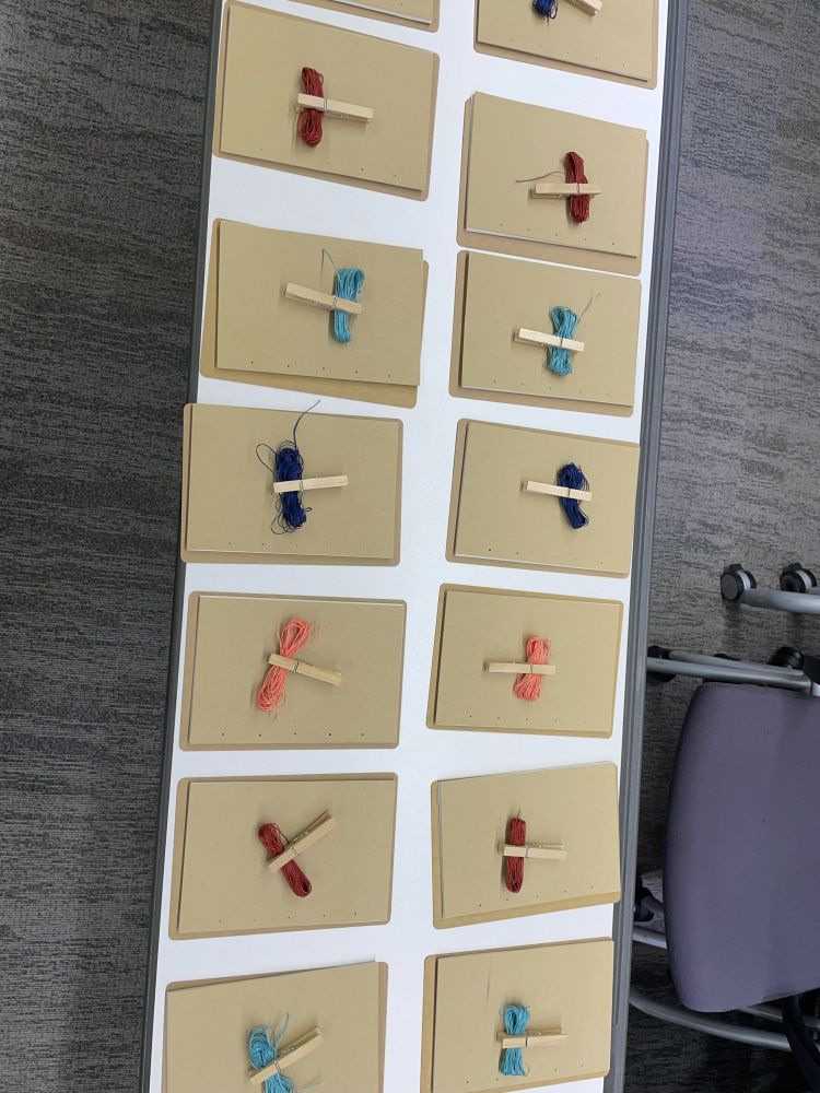 Thread binding notebook supplies laid out on a table with grey carpet and chairs in the background. Coloured bundles of thread are clipped together with clothespins on top of brownish looking sheafs of paper.
