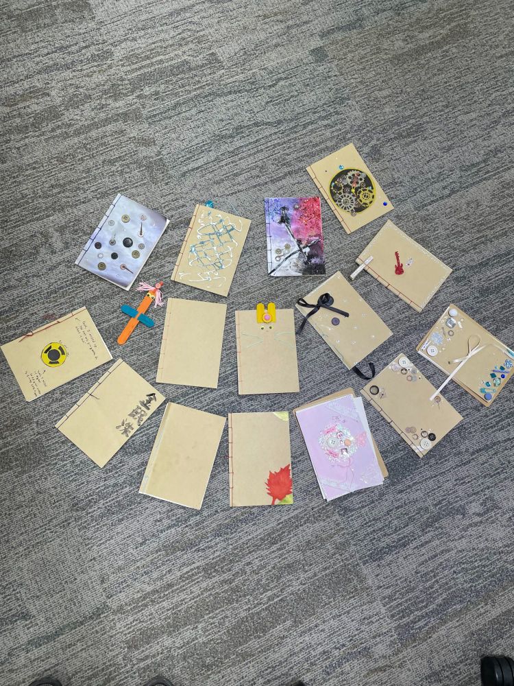 Fifteen thread-bound notebooks are spread out on a mottled grey commercial carpet. Each one is decorated in a different style, including hand-drawn art, characters, glued decorations, or glitter. There's also a character bookmark.