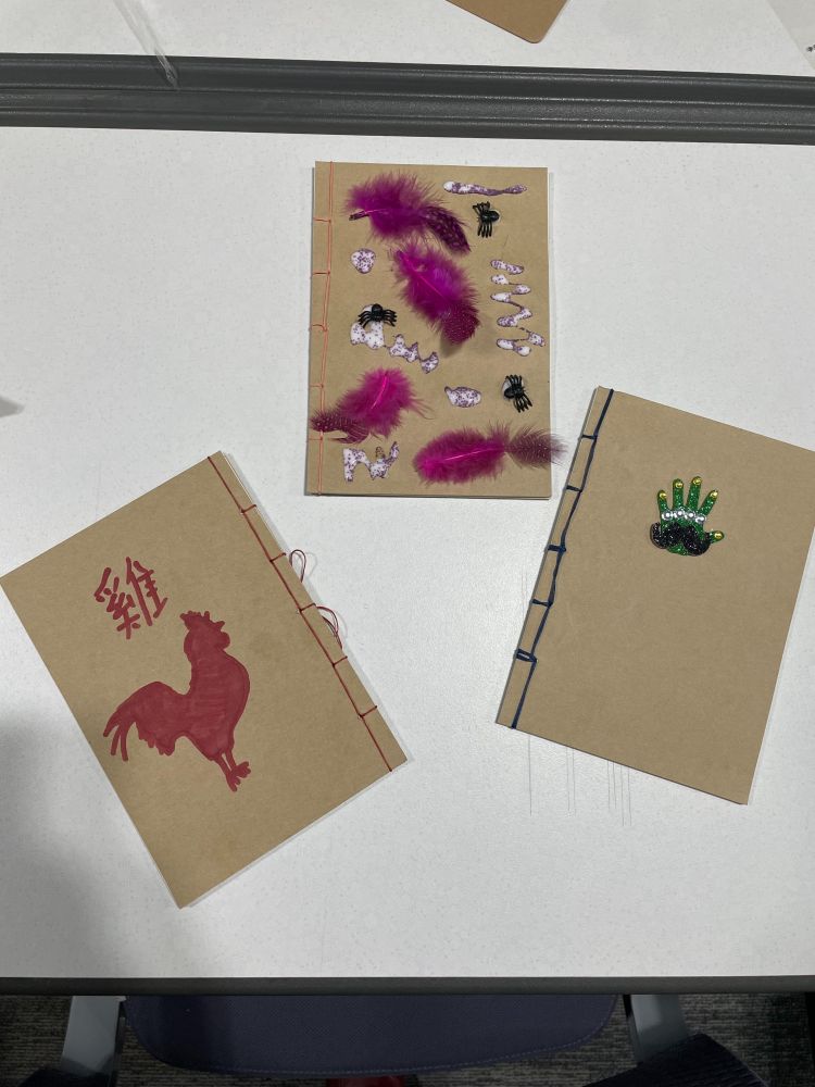 Three thread-bound notebooks with red, pink and dark green thread bindings and a red hand-drawn art, glue, fuschia feathers and toy spiders, and a plastic sparkly green hand on natural/brownish tone covers.