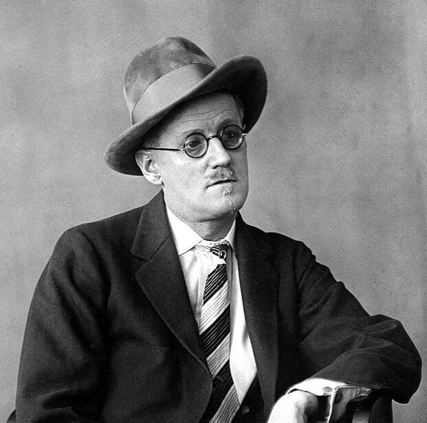 Black and white portrait of James Joyce seated on a wooden chair. He is wearing a suit, a wide-brimmed hat, round glasses, and a striped tie. He gazes slightly away from the camera with a composed expression.