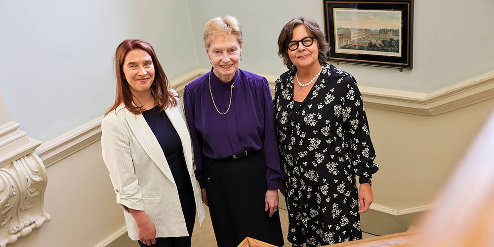 Nicole Grimes (left), Rosemary O’Mahony (centre), and Eve Patten (right) stand together on a staircase. Nicole wears a white blazer, Rosemary wears a purple blouse, and Eve wears a black floral dress. A framed artwork hangs on the wall behind them.