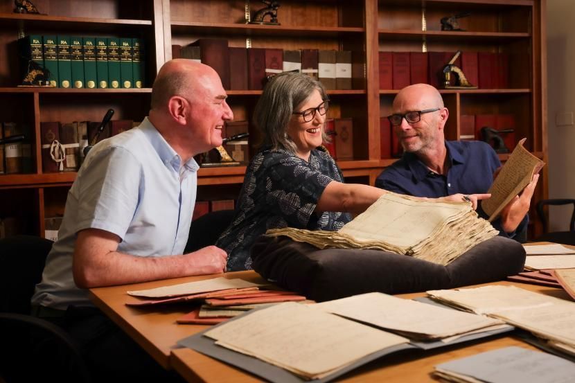 Three people smiling and examining historical documents at a table, surrounded by shelves of archival books and manuscripts. The atmosphere is warm and collaborative, highlighting the team effort behind the Virtual Record Treasury of Ireland.