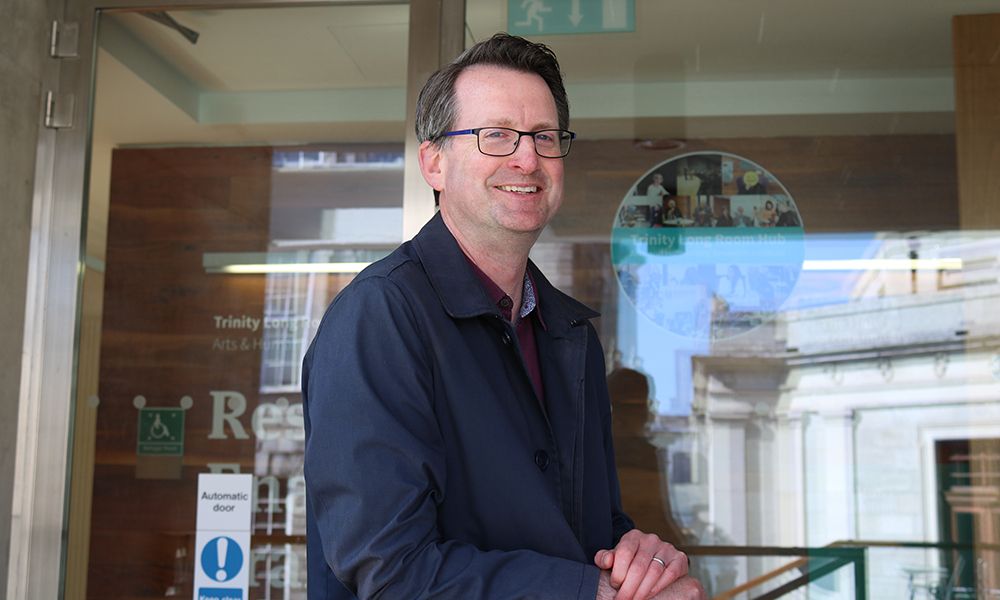 Professor Patrick Geoghegan standing outside the entrance to the Trinity Long Room Hub, smiling with hands clasped. The Hub logo and part of the Trinity College Dublin campus are visible in the background through the glass doors.