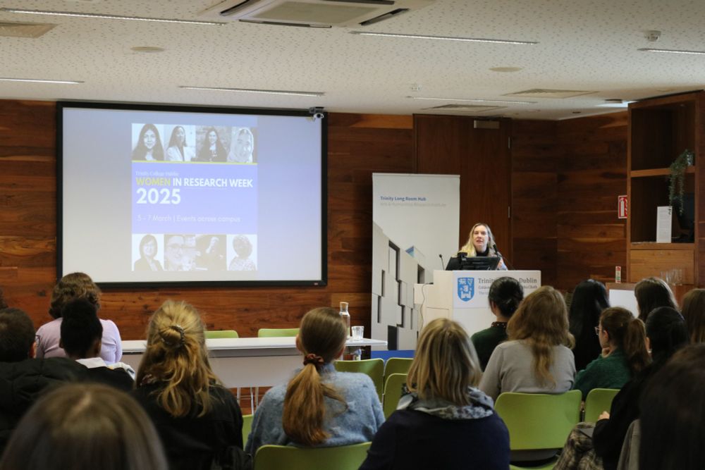 PhD researcher and conference organiser Morgiane Noel standing at a lectern speaking to an audience with a powerpoint presentation projected on a screen