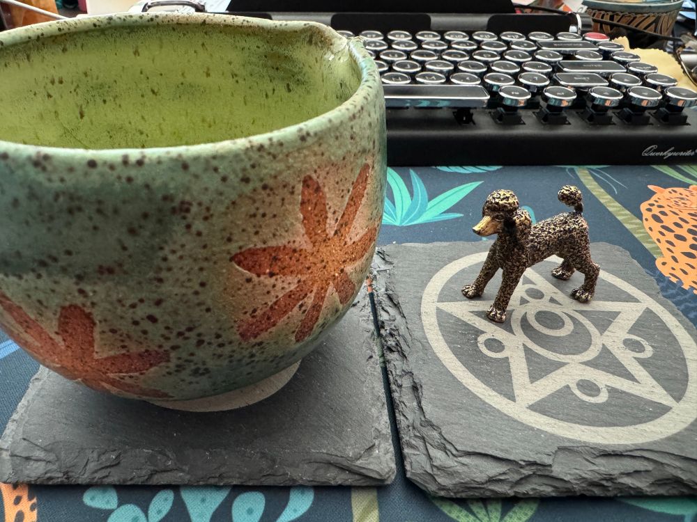 Green matcha chawan/cup with golden brown flower design and a small bronze poodle statue, both on gray slate coasters. 