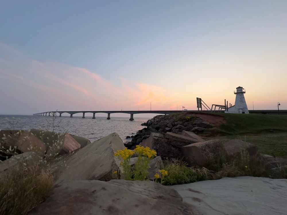 Confederation Bridge, PEI. Rocky shore in the foreground. Lighthouse on the right. Sunset behind. 
