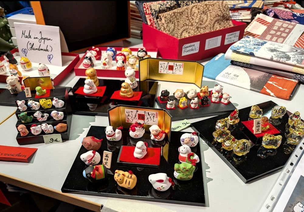 Ein Tisch auf dem schwarze Brettchen liegen und auf diesen werden kleine, rundliche, süße Tiere zum Kauf angeboten.
Z.b. die chinesischen Tierkreiszeichen und Winkekatzen.
Material wahrscheinlich Glas, Porzellan, Keramik.
Weiter hinten stehen und liegen schön gemusterte japanische Stoffe.
