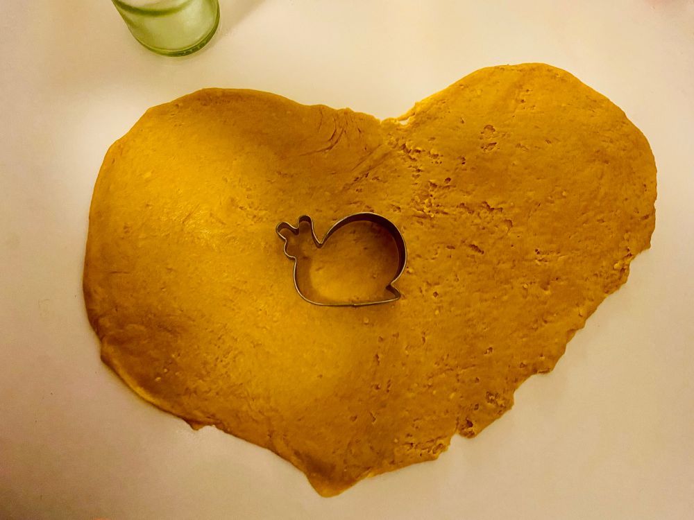 Ein in Herzform ausgerollter dunkler Teig auf einem weißen Tisch. Darauf liegt ein Backförmchen in Schneckenform.