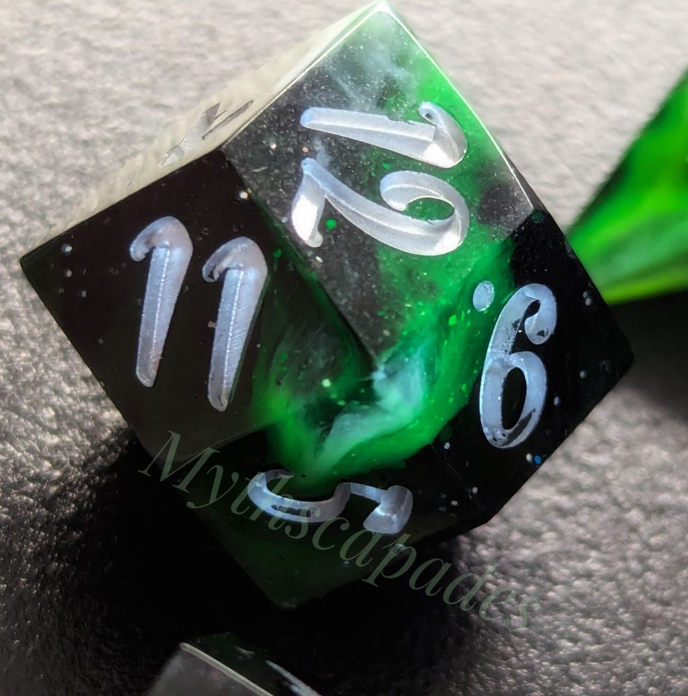 A closeup of the d12 of the set of roleplaying dice that are black transparent with silver sparkles and swirls of green and white looking like an aurora borealis. Inked in silver