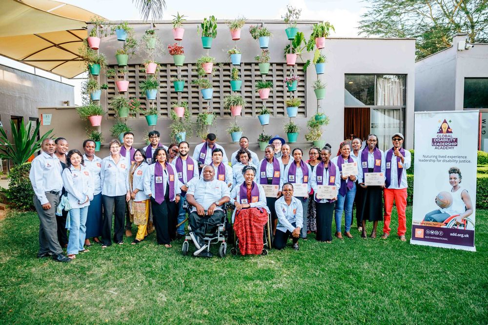 Global Disability Leadership Academy participants and facilitators stand together outdoors in front of a building with a colorful vertical garden made of hanging pots. Everyone is smiling, some holding certificates, and many raising their fists in celebration. Several participants wear purple stoles over white shirts. There is a banner with the Global Disability Leadership Academy branding and the tagline “Nurturing lived experience leadership for disability justice.”