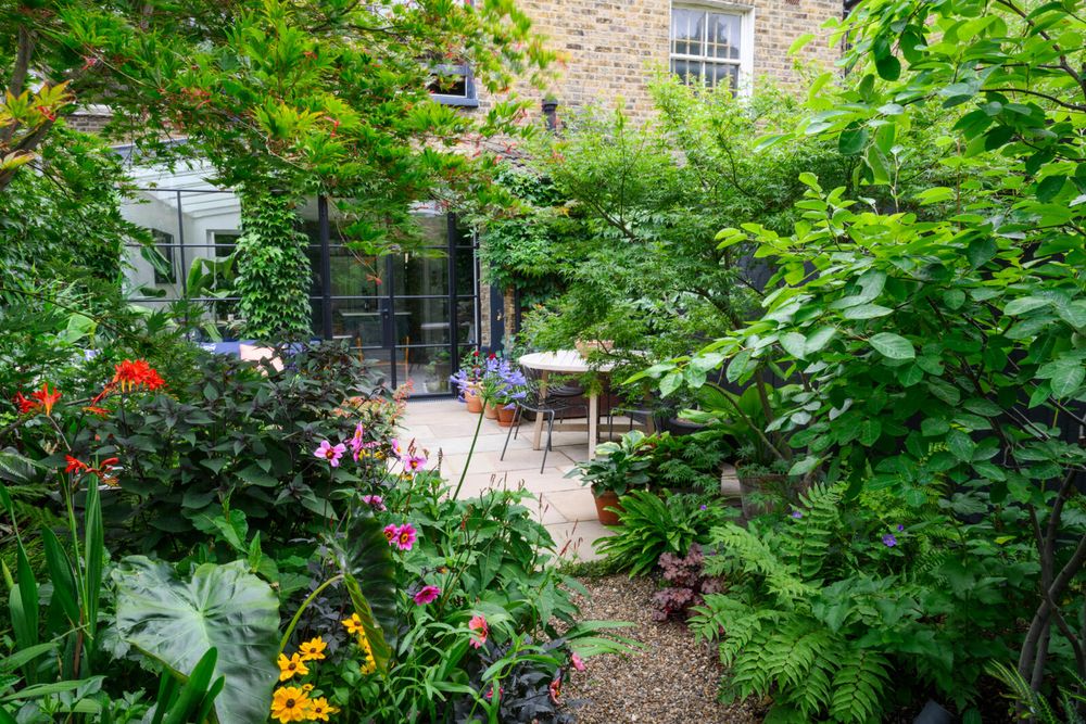 A lush garden in front of a stone building with large glass doors, featuring diverse plants, flowers in bloom, and a wooden table set.