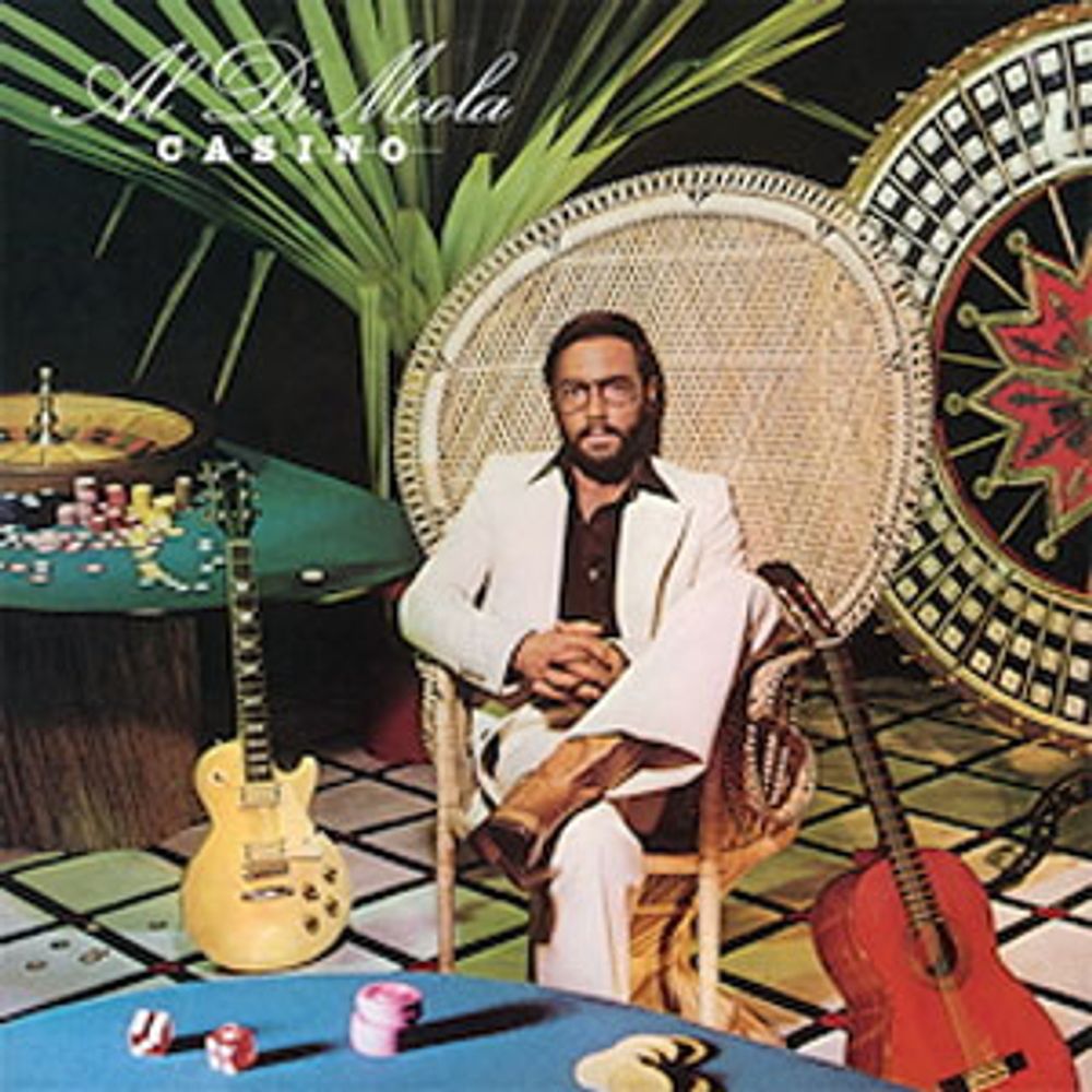 Al Di Meola seated in an ugly chair near a casino table between a Les Paul goldtop and an acoustic guitar.