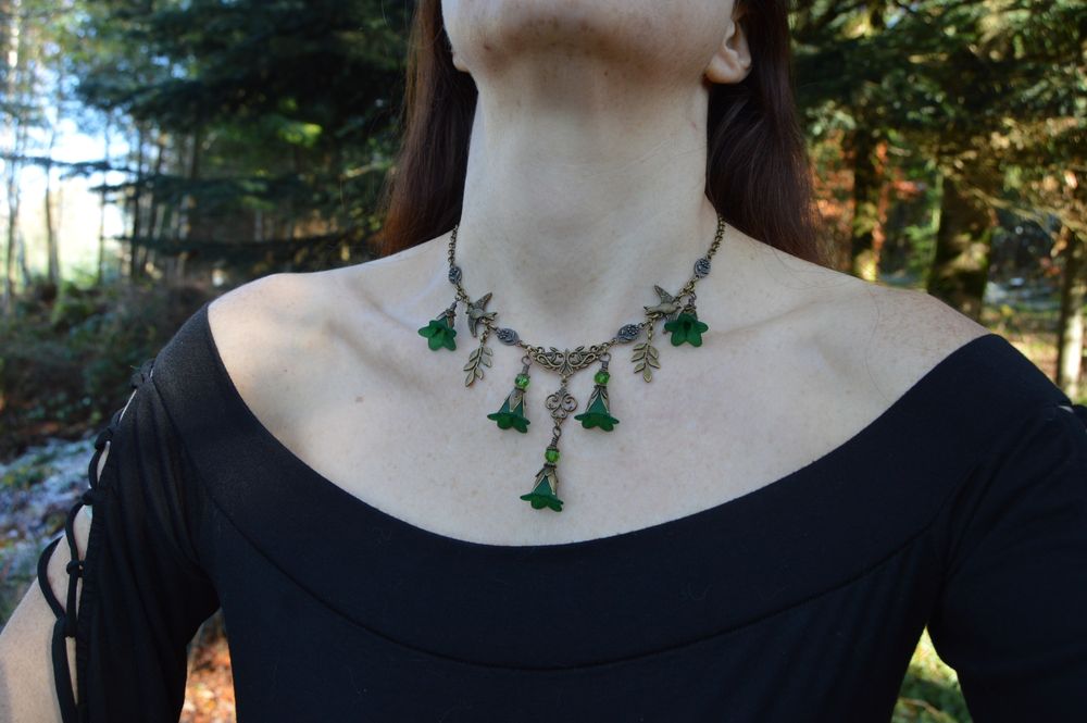 Unedited photo of a bronze necklace with dark green flowers, worn by a pale figure in a black dress standing in a forest. Frost on the ground and sunlight through dark pine trees. The necklace is v shaped, with 5 dangling flower pendants in dark emerald green. Small leaf, flower and bird charms dot the chain.