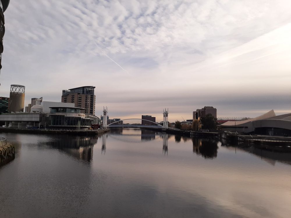 Footbridge going across the Manchester Ship Canal. Waterfront buildings on either side, mottled clouds in the sky and the water is still. A cold winter day.