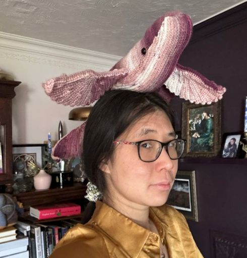 Award-winning Author and crafty crochet-er, Jeannette Ng, with dark hair, middle parting, black glasses, giving a cute side eye, wearing a silky mustard yellow shirt, with a bespoke crocheted beasts resting atop their head - a pink and purple whale with winged fins. Bakground has a collection of books, interesting paintings. 