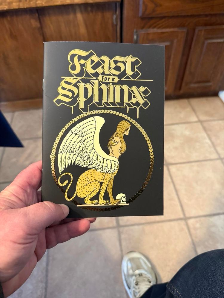 A hand holds a black booklet titled "Feast for a Sphinx," featuring a golden illustration of a sphinx with wings in the center. The background shows a tiled floor and a wooden surface.