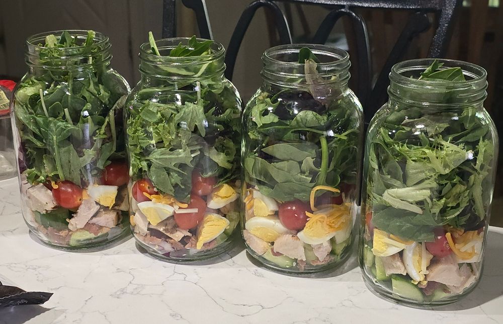 4 Mason jars are filled with chopped veggies, grilled chicken, shredded cheese, bacon pieces, and mixed greens.