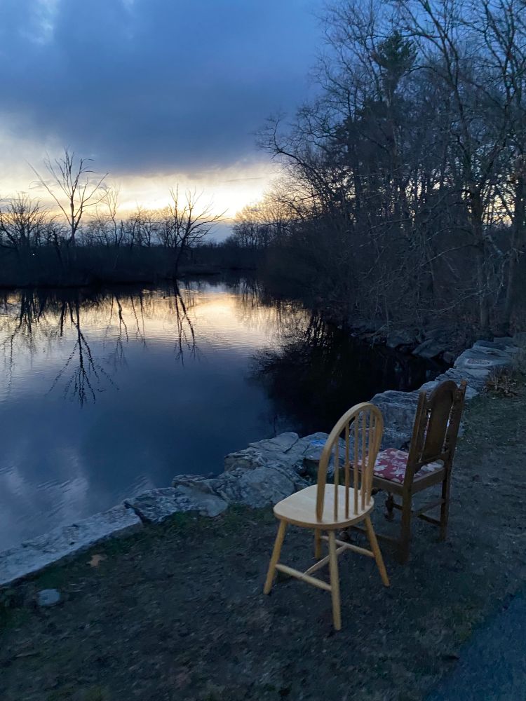 Two chairs are in the grass by the side of the paved running path. They are facing away, looking out over a small pond, with barren trees all around. It’s early sunset with some yellow rays still peeking through, but there are some dark clouds covering a good portion of the sky.