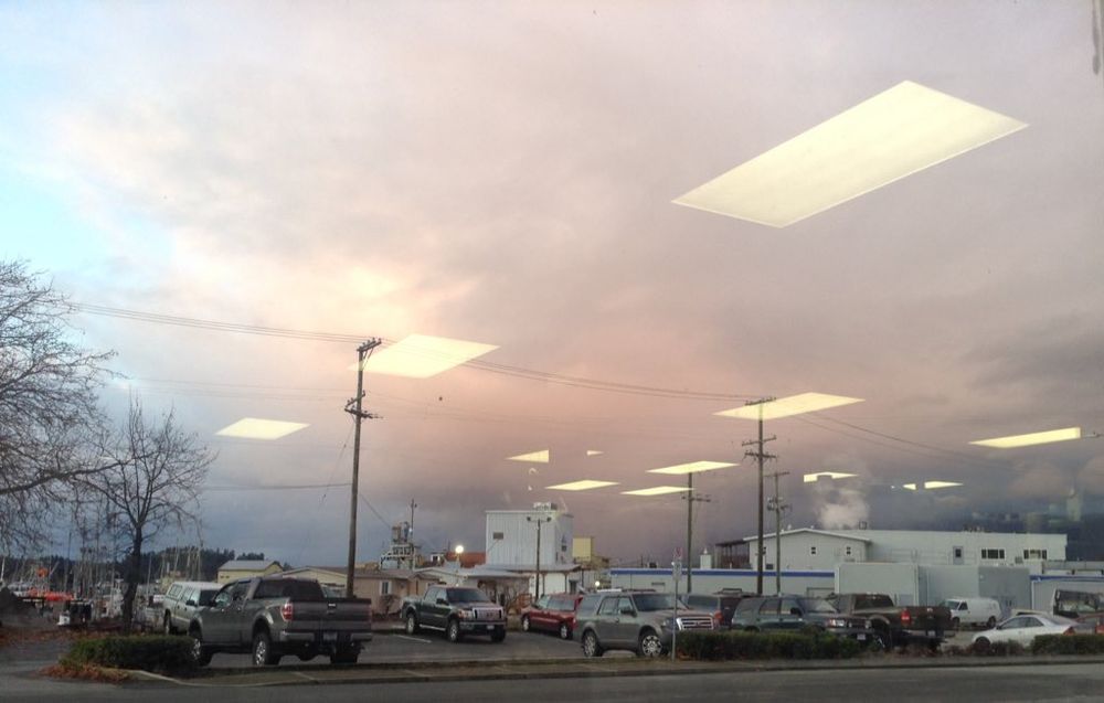  photo of a parking lot with a few vehicles, behind which are a couple of white industrial-looking buildings (ice plant / fish processing) and a gathering storm cloud which is tinted pink from the setting winter sun. There also appear to be a bunch of 24x48 inch fluorescent ceiling light panels, which are actually the reflection of the ceiling of the store in which i took this photo