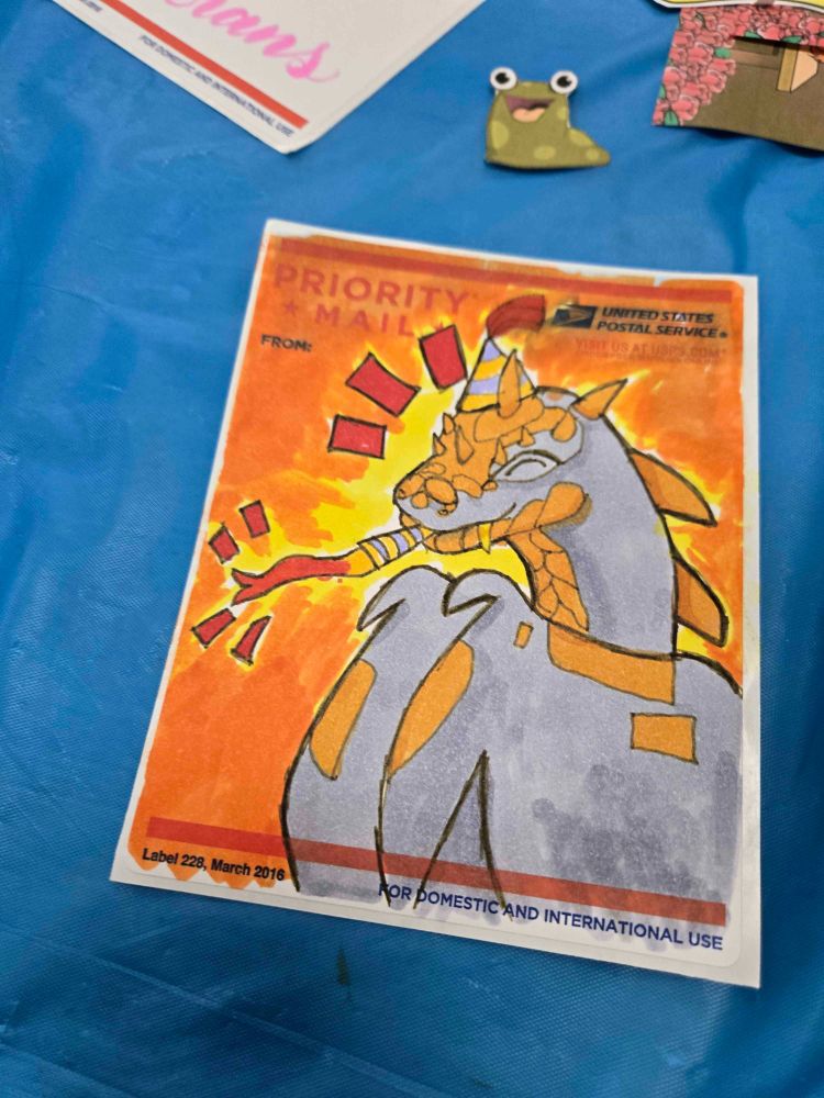 a picture of a drawing of a tyrannian Hissi with a party hat and noisemaker with a yellow and orange background the drawing is on a blue table with multiple pieces of paper including a slorg