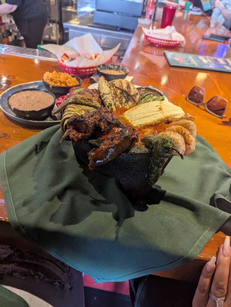 Stone molcajete filled with carne asada, pollo asada, shrimp, nopales, jalapenos, and cheese, on a napkin at a bar 