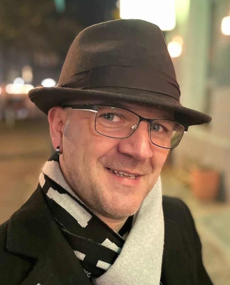 Portraitfoto von Marcel. Er lächelt direkt in die Kamera. Er trägt einen Hut und eine Brille. Dazu einen schwarz-weißen Schal und einen schwarzen Mantel.