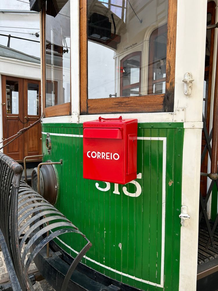 an old tram in a museum. the tram is green with white trim, and has a black grille-cum-cow catcher on the front. on the tram is a bright red mailbox that says CORREIO, the portuguese word for mail