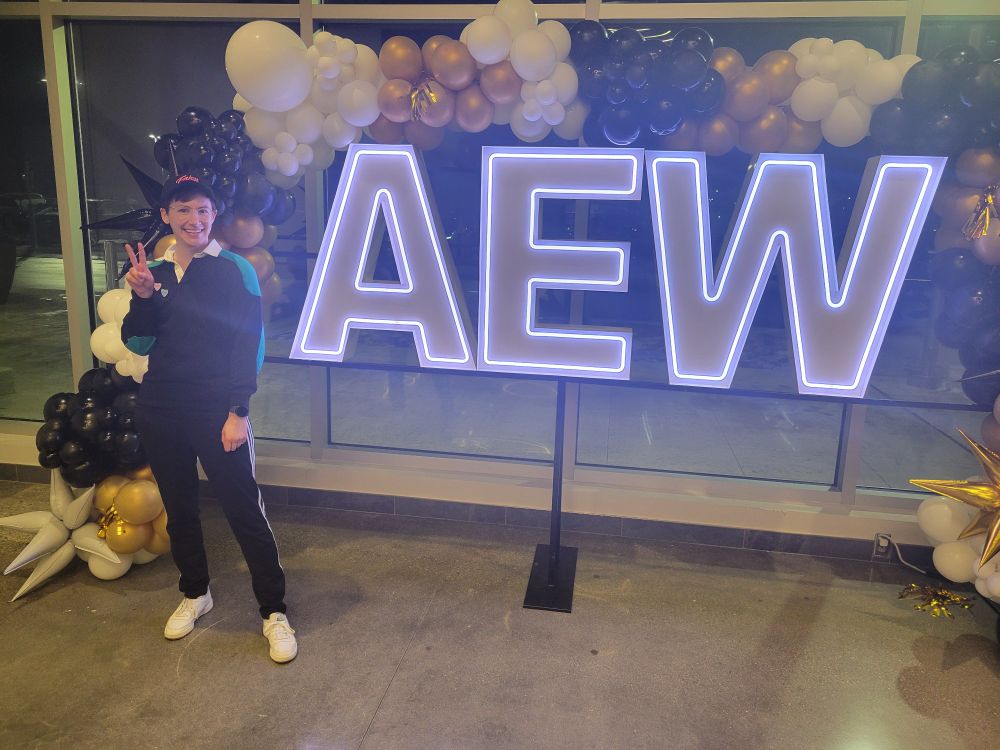 me looking cute in front of the neon aew sign with gold black and white balloon arch