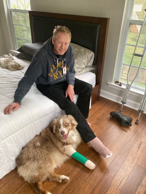 An older man sits on a bed with a slight smile and his right ankle in a cast. A dog lays beside him with a bright expression on, and a front right leg in a cast. Apparently the dog injured its leg hopping onto a boat and the dad injured his ankle when he went to go check on it.