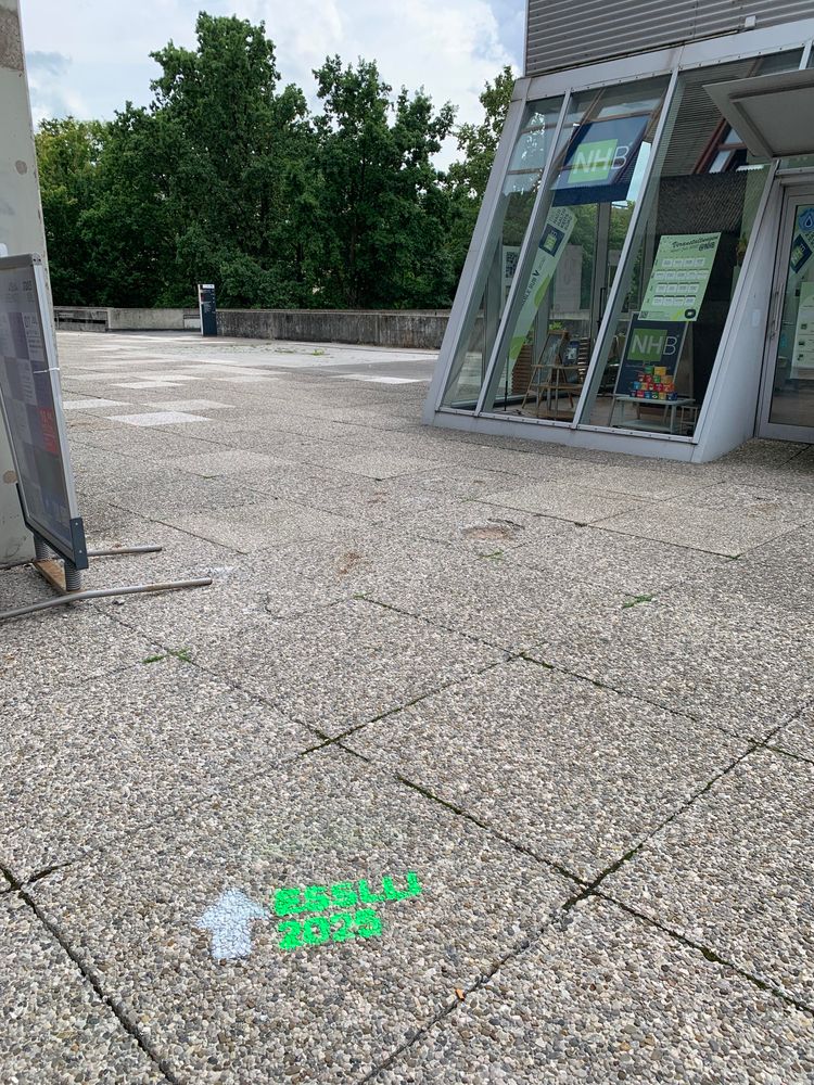 Concrete path with  “ESSLLI 2025” and a large arrow sprayed in green and white chalk. A building can be seen on the side and some trees in the background 