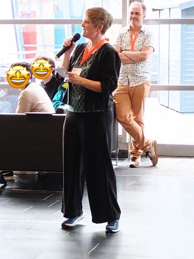 Woman (me) in black clothes standing on tip-toes and speaking into a microphone.  Behind me, program chair Balder ten Cate has his arms crossed and is smiling. 