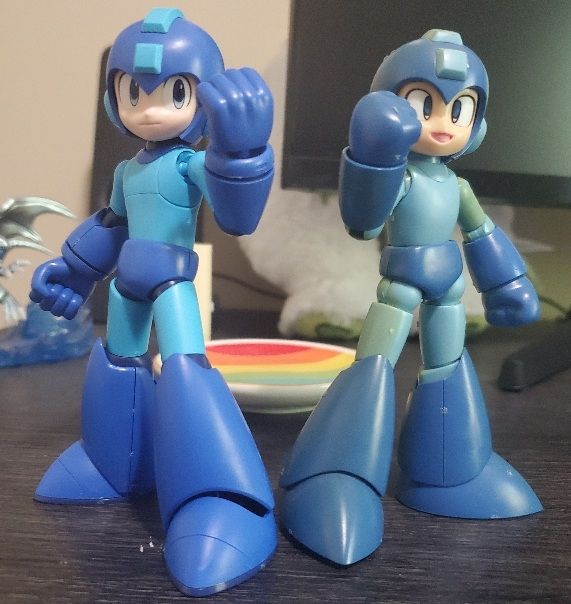 Two Mega Man model kits standing side by side, each with a fist raised towards the camera triumphantly. The one on the left is the newer model with mode detail and the one on the right is the older model with less detail and its colors are faded and yellowed with age. 