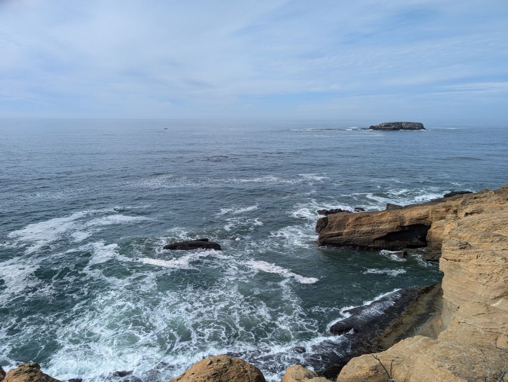 A picture of the ocean with more waves in it. Taken near the Devil's Punchbowl probably. There's an outcrop of dark stone in the distance, but mostly just ocean and sky. The ocean here looks greenish blue.