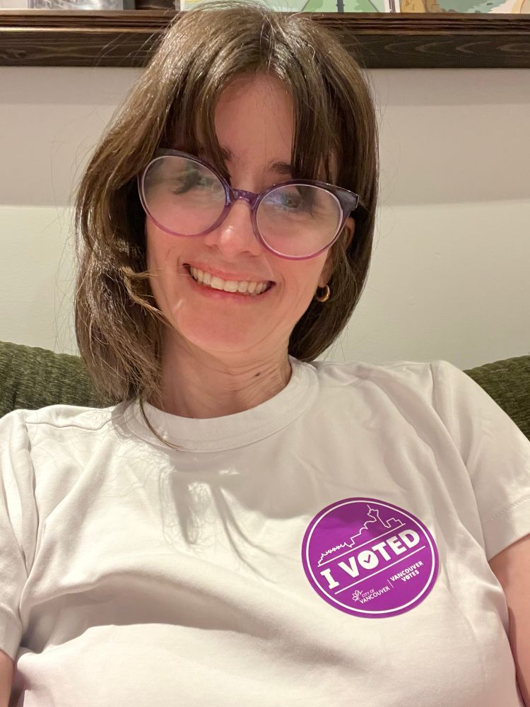 A brown haired middle-aged woman smiling. She has a purple “I voted” sticker on her white shirt. 