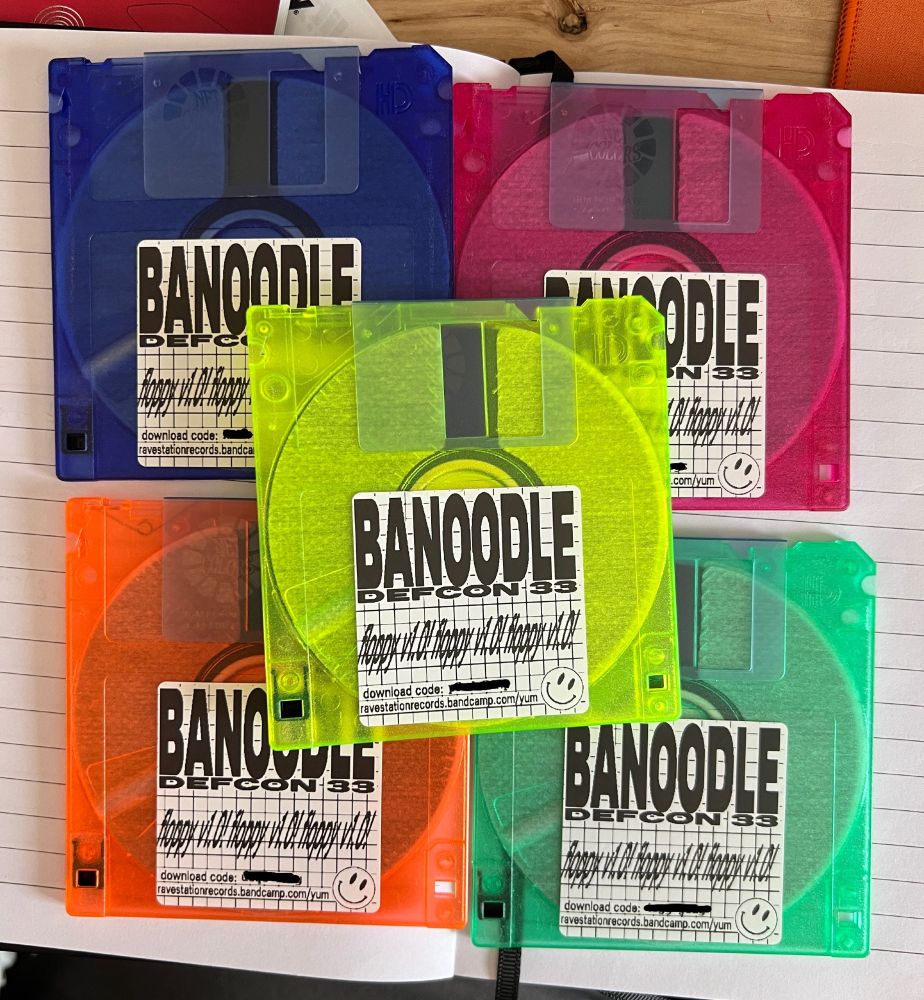 A stack of five neon-colored clear floppy disks, labeled "BANOODLE - DEFCON 33 - floppy v1.0!". At the bottom, they have a Bandcamp download code and a link to the RaveStation Records Bandcamp page, where the code can be redeemed.