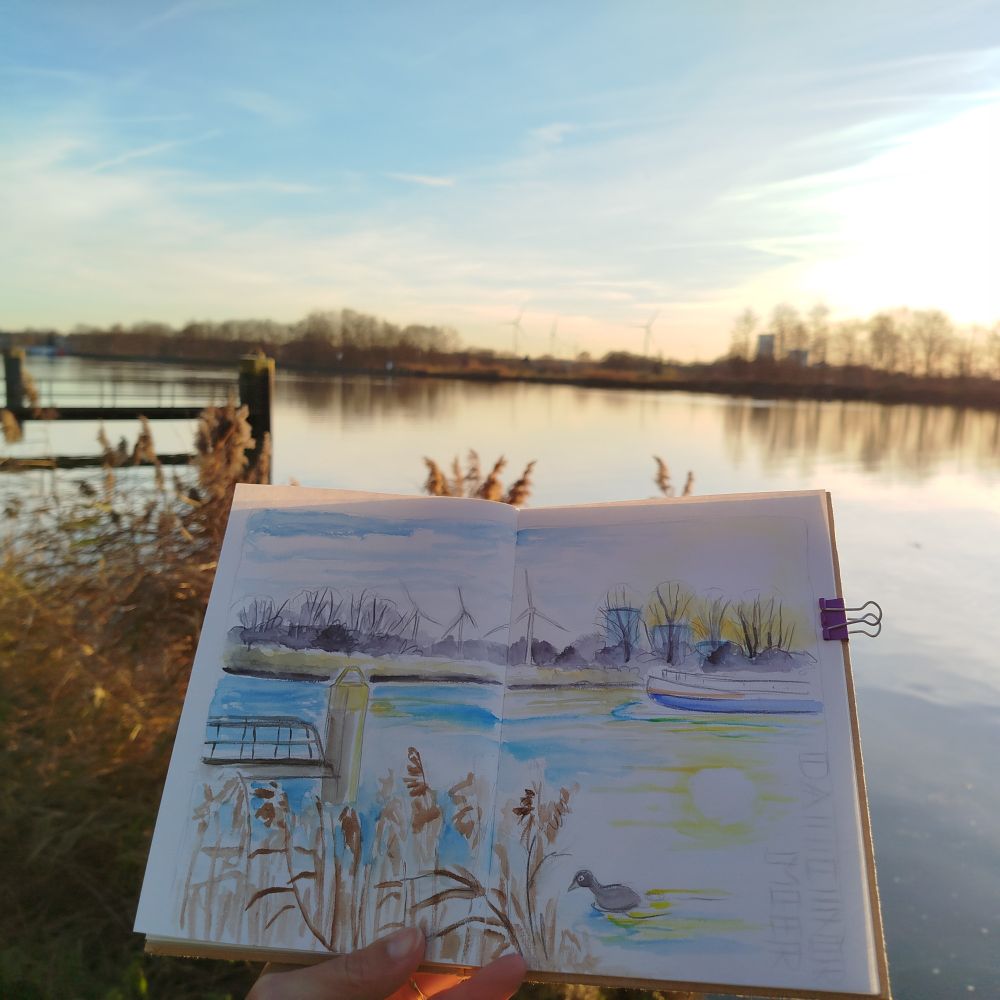 Foto des Skizzenbuchs vor einer großen Wasserfläche. Im Vordergrund vertrocknetes Schilf, Steganlage, im Hintergrund Uferlinie mit winterkahlen Bäumen, Windrädern, Kraftwerk. Blauer Himmel mit Wolkenschlieren und tiefstehende Sonne.