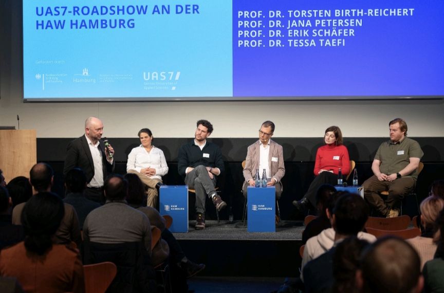 fünf Personen sitzen auf einem Podium und hören einer stehenden Person im Publikum zu